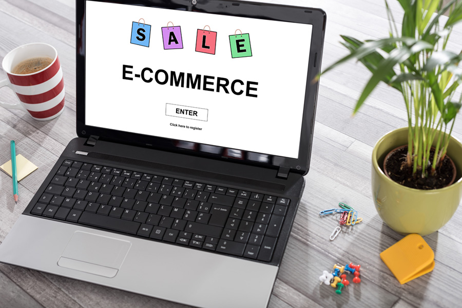 the words Sale and ECommerce on a laptop screen with an enter button. There is a plant on the right side of laptop and a cup of coffee on the left side.